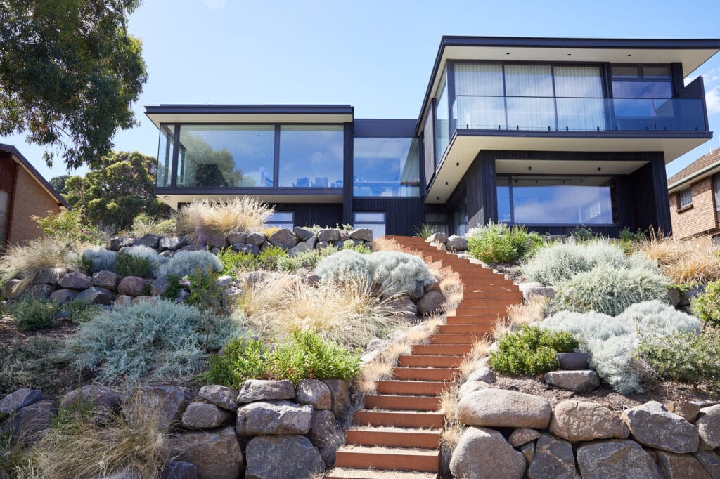 A modern, two-storey house with large glass windows sits atop a hill, with stone steps and landscaped plants leading up through rocky terrain. Loci Landscapes | Landscape Designers Tasmania