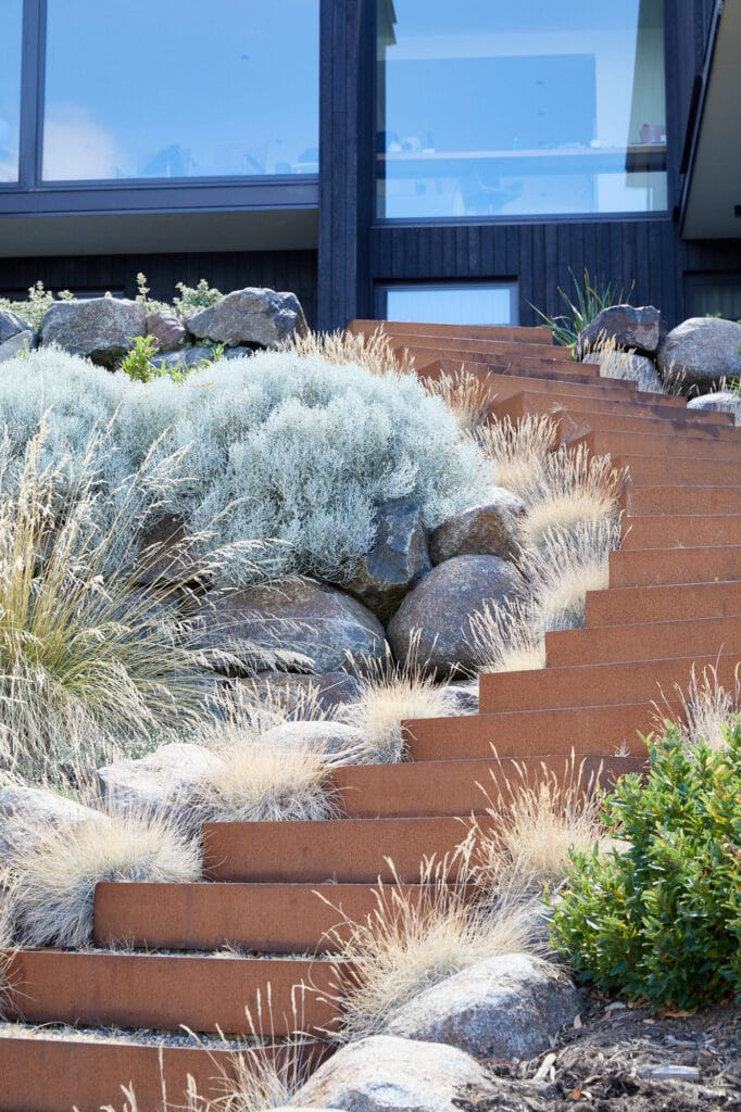 Rust-coloured metal steps ascend through ornamental grasses and rocks, leading to a modern building with dark cladding and large windows in the background. Loci Landscapes | Landscape Designers Tasmania