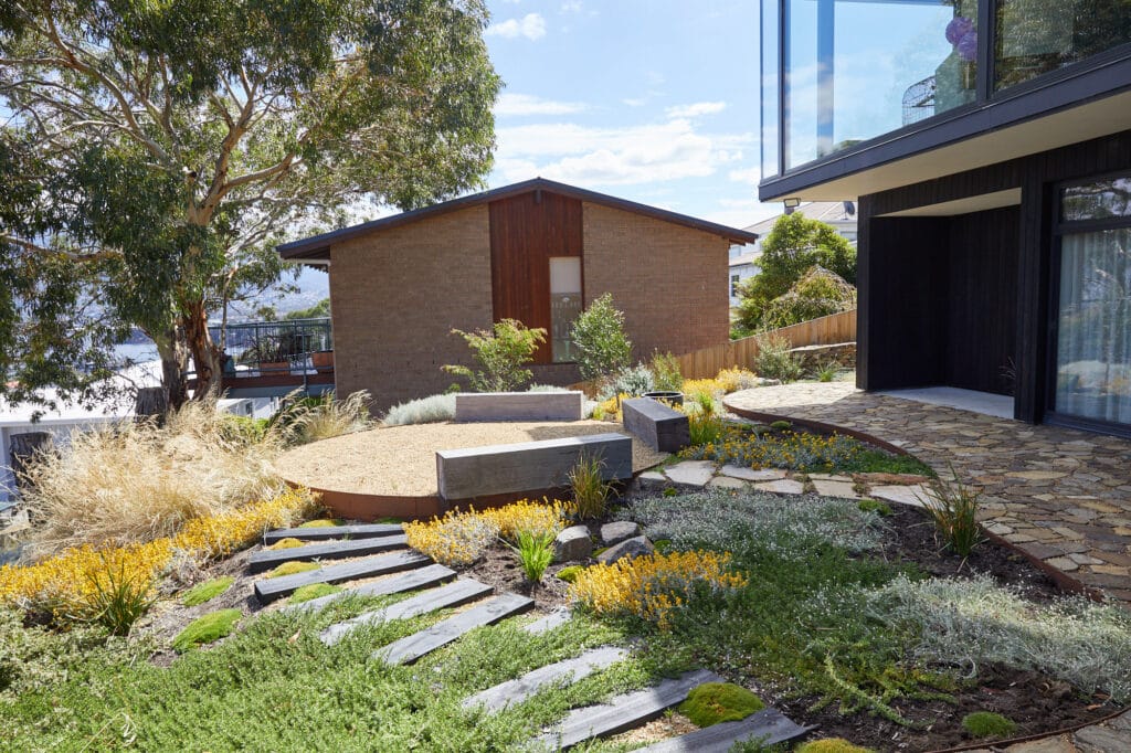 Modern landscaped garden with stone paths, raised circular seating area, wooden benches, and a mix of grasses and shrubs; a house and outbuilding are visible in the background. Loci Landscapes | Landscape Designers Tasmania