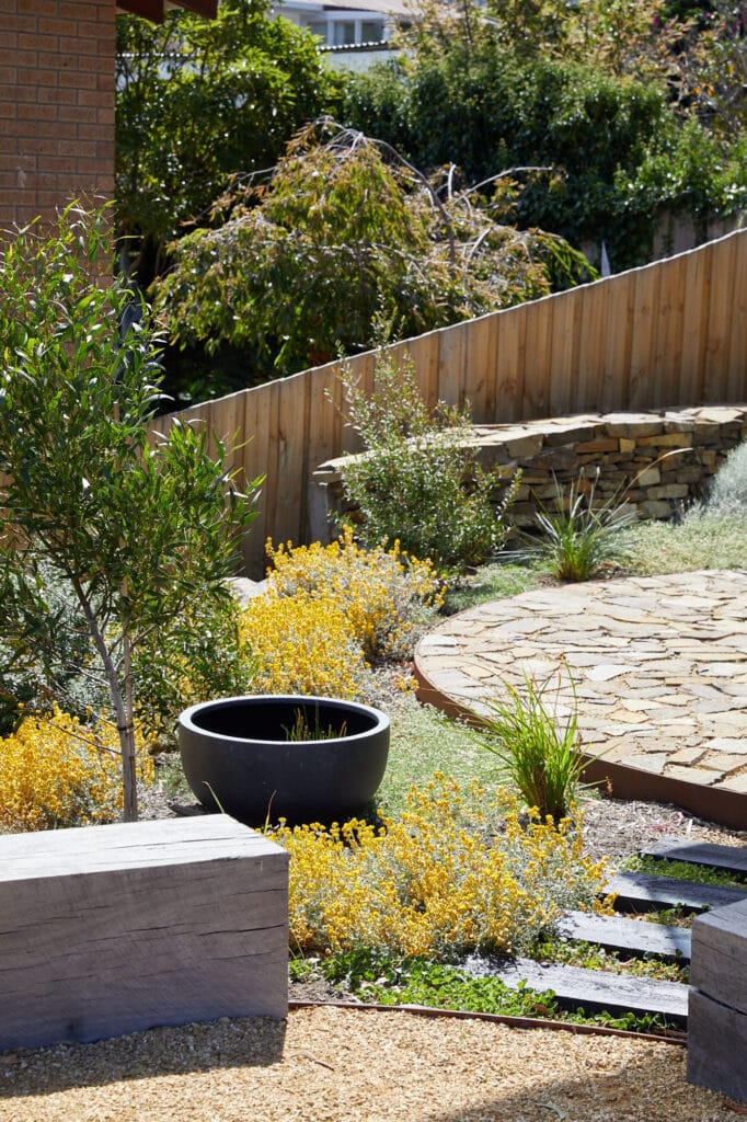 A landscaped back garden with a stone path, yellow flowering plants, a black planter, wooden fence, and stone bench. Loci Landscapes | Landscape Designers Tasmania