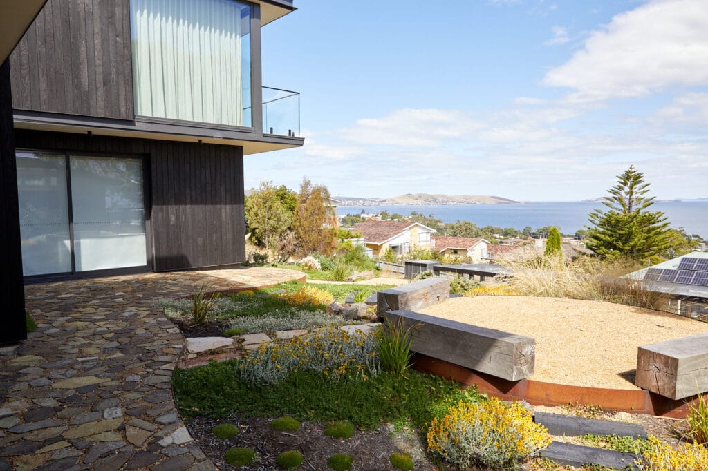 Modern house with stone patio, large windows, and landscaped garden overlooking a coastal neighbourhood and the sea under a partly cloudy sky. Loci Landscapes | Landscape Designers Tasmania