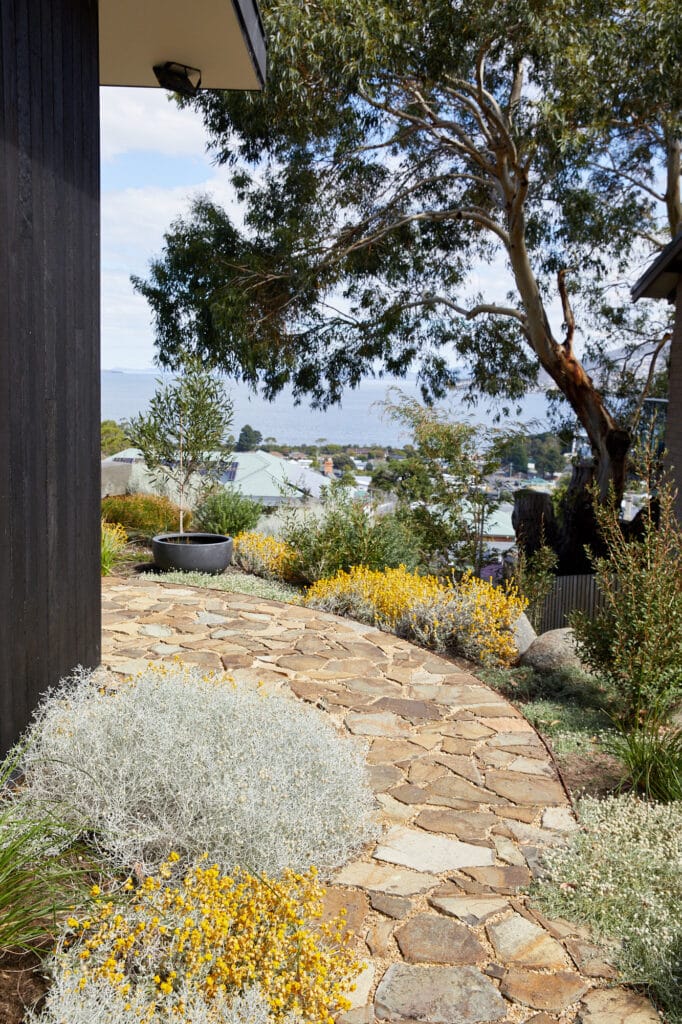Curved stone path bordered by shrubs and yellow flowers, with a view of rooftops, trees, and distant water in the background. Loci Landscapes | Landscape Designers Tasmania