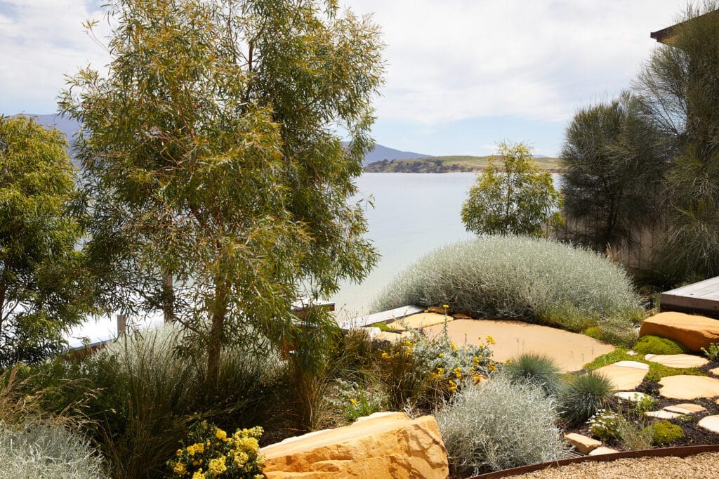 A garden with native plants and large rocks overlooks a calm lake, with distant hills and a partly cloudy sky in the background. Loci Landscapes | Landscape Designers Tasmania