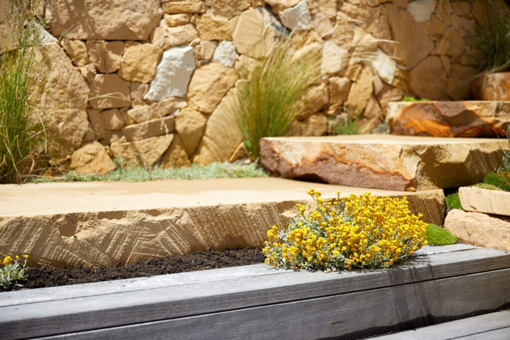 Yellow flowers grow beside large stone steps and a wooden planter, with a stone wall and decorative grasses in the background. Loci Landscapes | Landscape Designers Tasmania