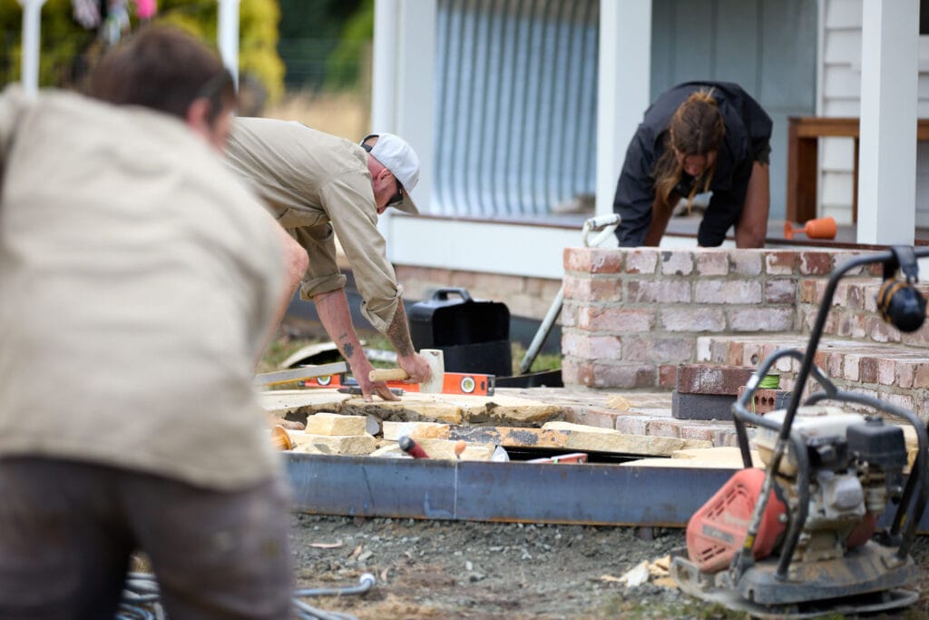 Three people work on a construction project outdoors, using tools to measure and lay bricks and stones near a partially built brick structure. Loci Landscapes | Landscape Designers Tasmania