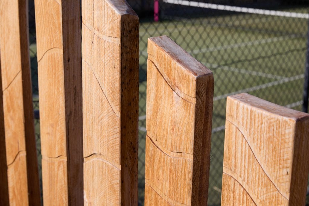 Close-up of several vertical wooden planks with carved patterns; a tennis court and chain-link fence are visible in the background. Loci Landscapes | Landscape Designers Tasmania