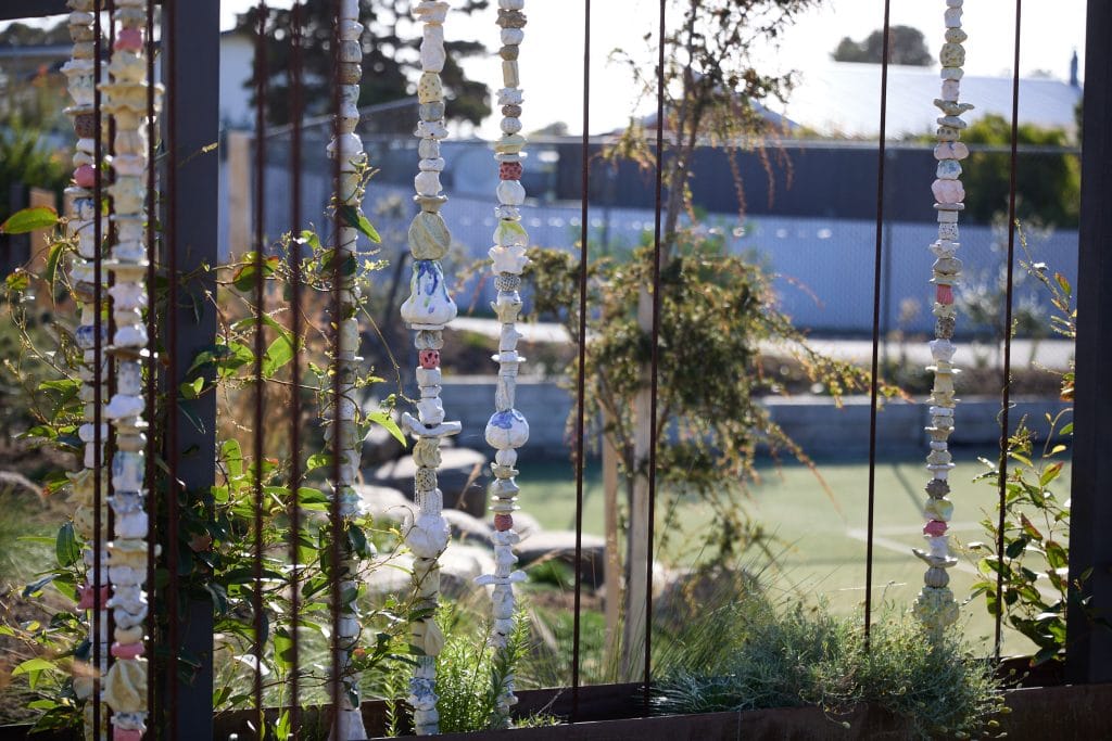 Vertical strands of colourful ceramic beads hang between metal bars in a garden with greenery and a blurred background of buildings and trees. Loci Landscapes | Landscape Designers Tasmania