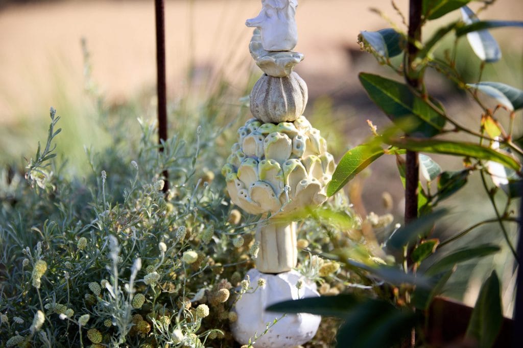 Ceramic garden ornament with stacked sculpted shapes stands among green foliage and small white flowers in a sunlit outdoor setting. Loci Landscapes | Landscape Designers Tasmania