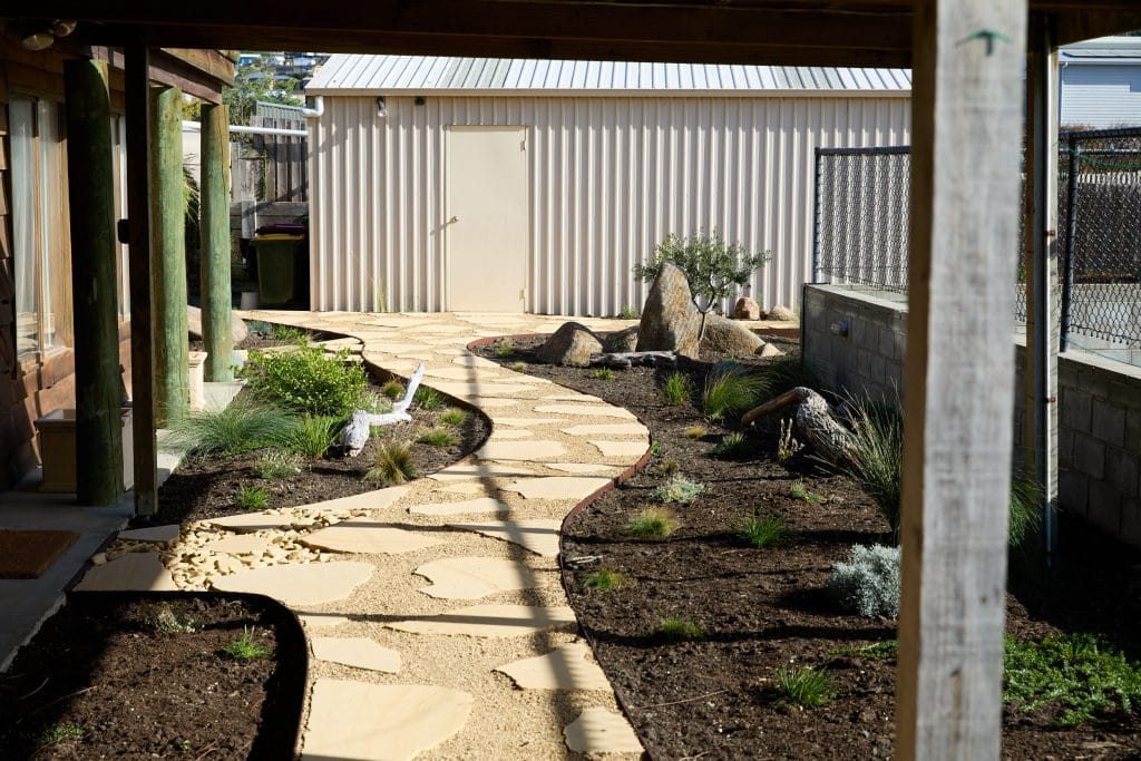 A back garden Native Garden with a winding stone pathway, various plants, rocks, and mulch, leading to a closed metal door on a corrugated shed—perfect for enjoying the charm of Primrose Sands. Loci Landscapes | Landscape Designers Tasmania
