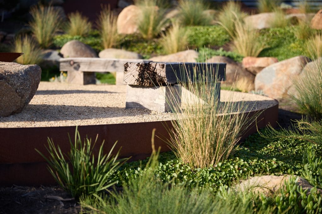A wooden bench sits on a gravel surface surrounded by ornamental grasses, rocks, and green plants in a landscaped garden. Loci Landscapes | Landscape Designers Tasmania