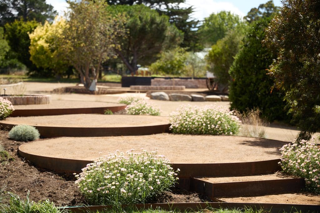 Circular raised garden beds with gravel surfaces are surrounded by flowering plants and trees in an outdoor landscaped area. Loci Landscapes | Landscape Designers Tasmania