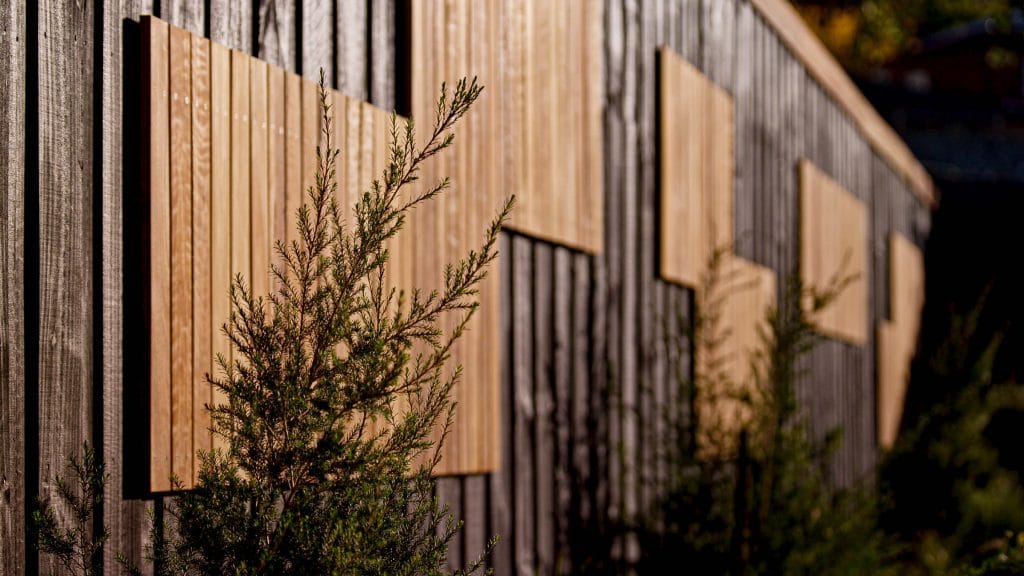 Close-up of a wooden fence with several rectangular wooden panels attached, partially obscured by green shrubs in the foreground. Loci Landscapes | Landscape Designers Tasmania