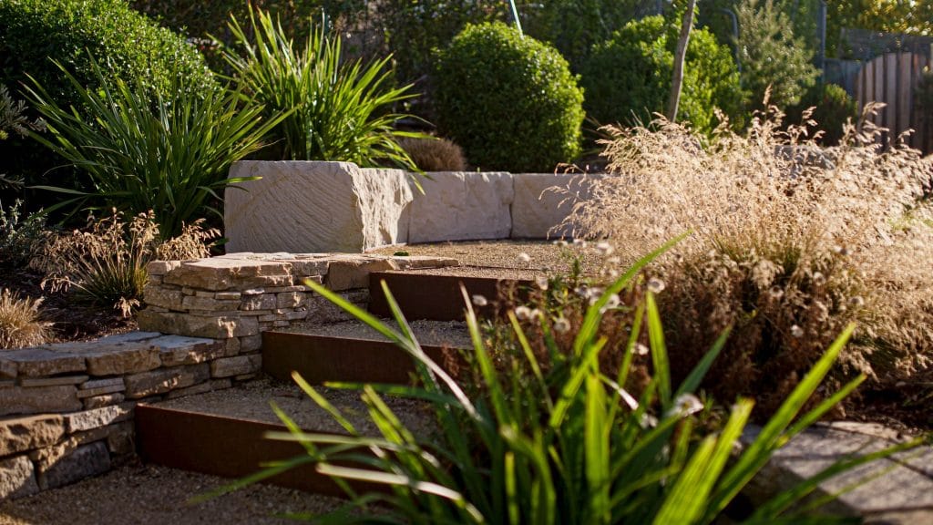 Stone steps lead through a landscaped garden with green shrubs, ornamental grasses, and a low stone wall in the background. Loci Landscapes | Landscape Designers Tasmania