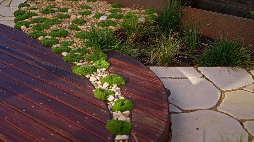 A curved wooden decking borders a landscaped area with gravel, round stepping stones, green moss, and ornamental grass. Loci Landscapes | Landscape Designers Tasmania