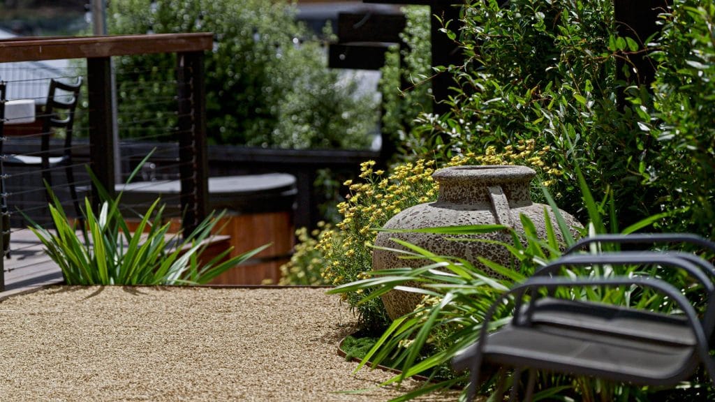 A gravel path with metal chairs, a large stone urn, green plants, and a wooden decking creates an inviting Small Garden Kingston setting. Loci Landscapes | Landscape Designers Tasmania